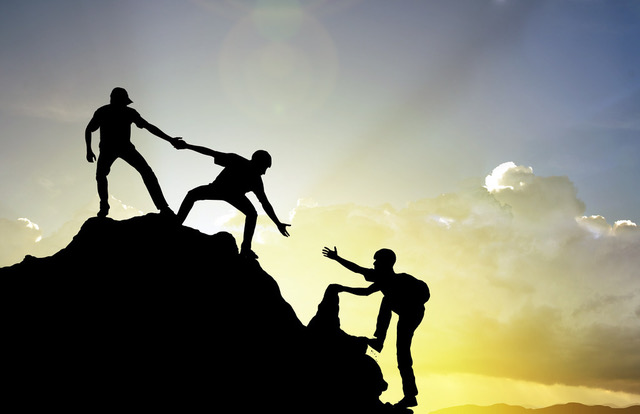 
	A group of three people hiking while two of them helps the last one climbing the top, with the sun behind them shining on them
